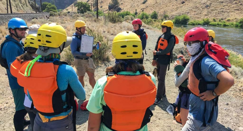 a group of students wearing life jackets and helmets watch as an instructor gives a rafting lesson
